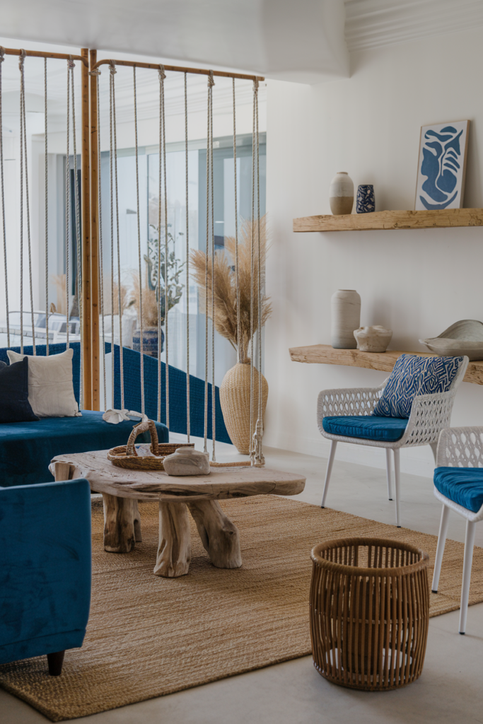 Modern living room with blue sofa, wooden coffee table, wicker chair, and decor items on shelves. Neutral tones with plants and pampas grass. Natural light fills the space.