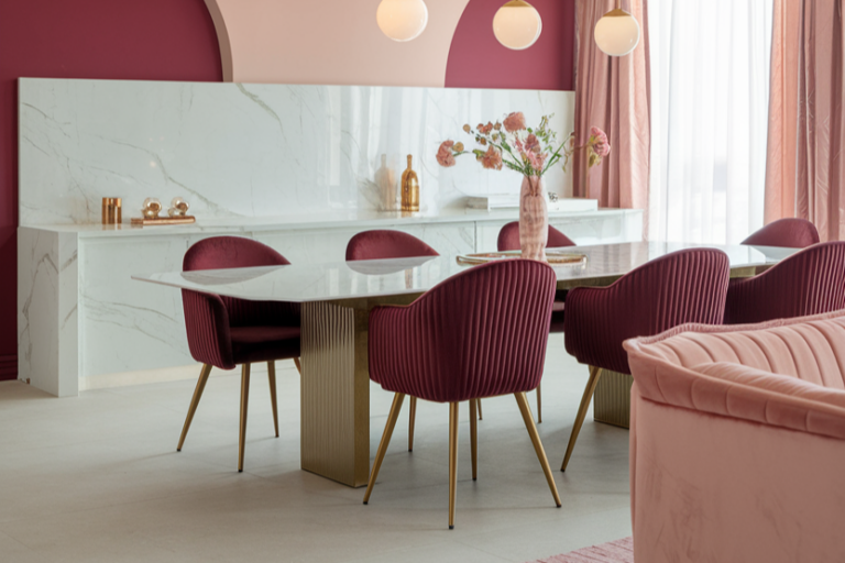 Dining area with a marble table, six red upholstered chairs, and a matching countertop. Pink and white walls, hanging lights, and a vase with flowers decorate the space.